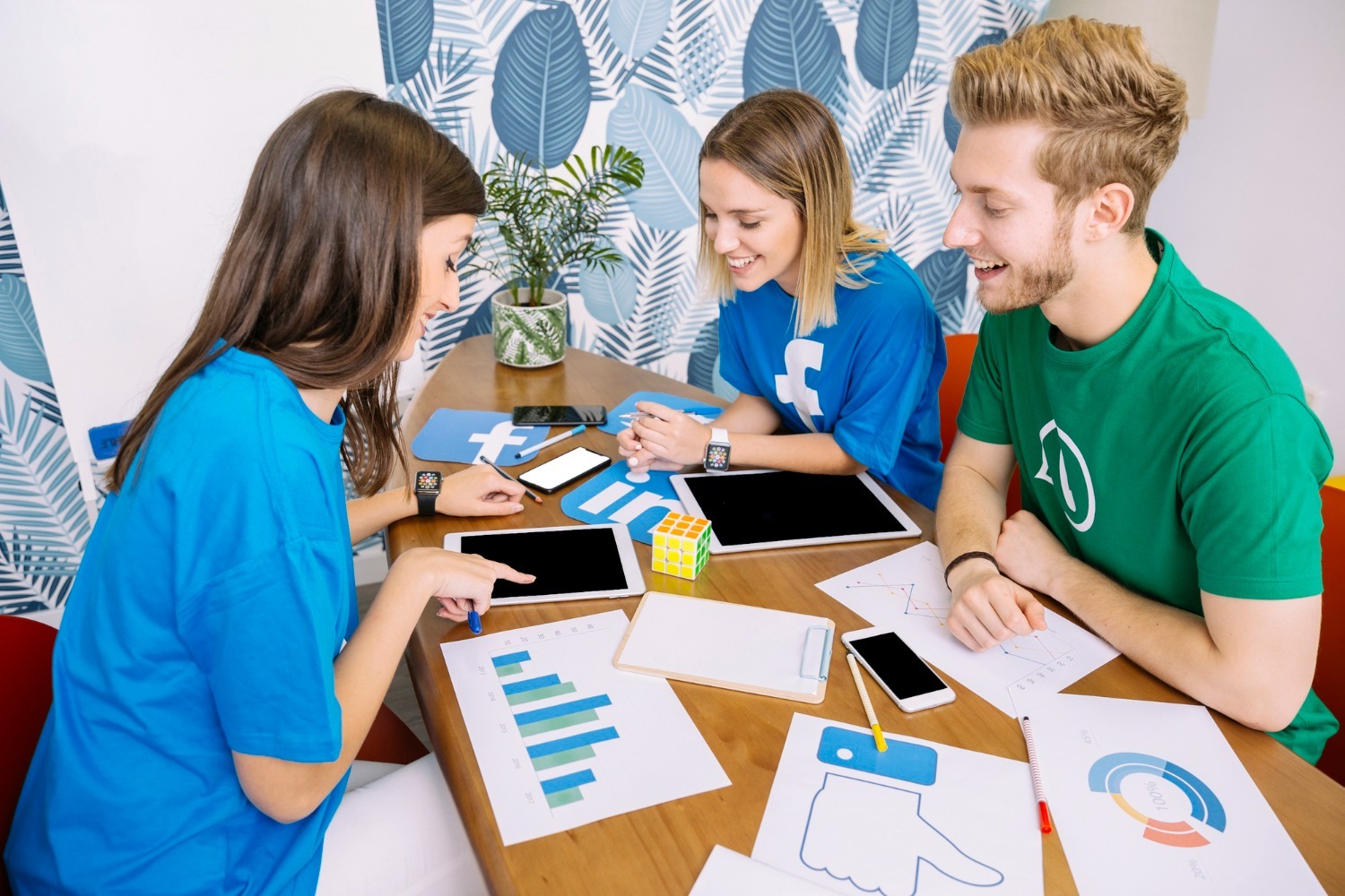 Three creative marketing professionals collaborating on a Facebook growth strategy with digital tablets, data charts, and social media branding.